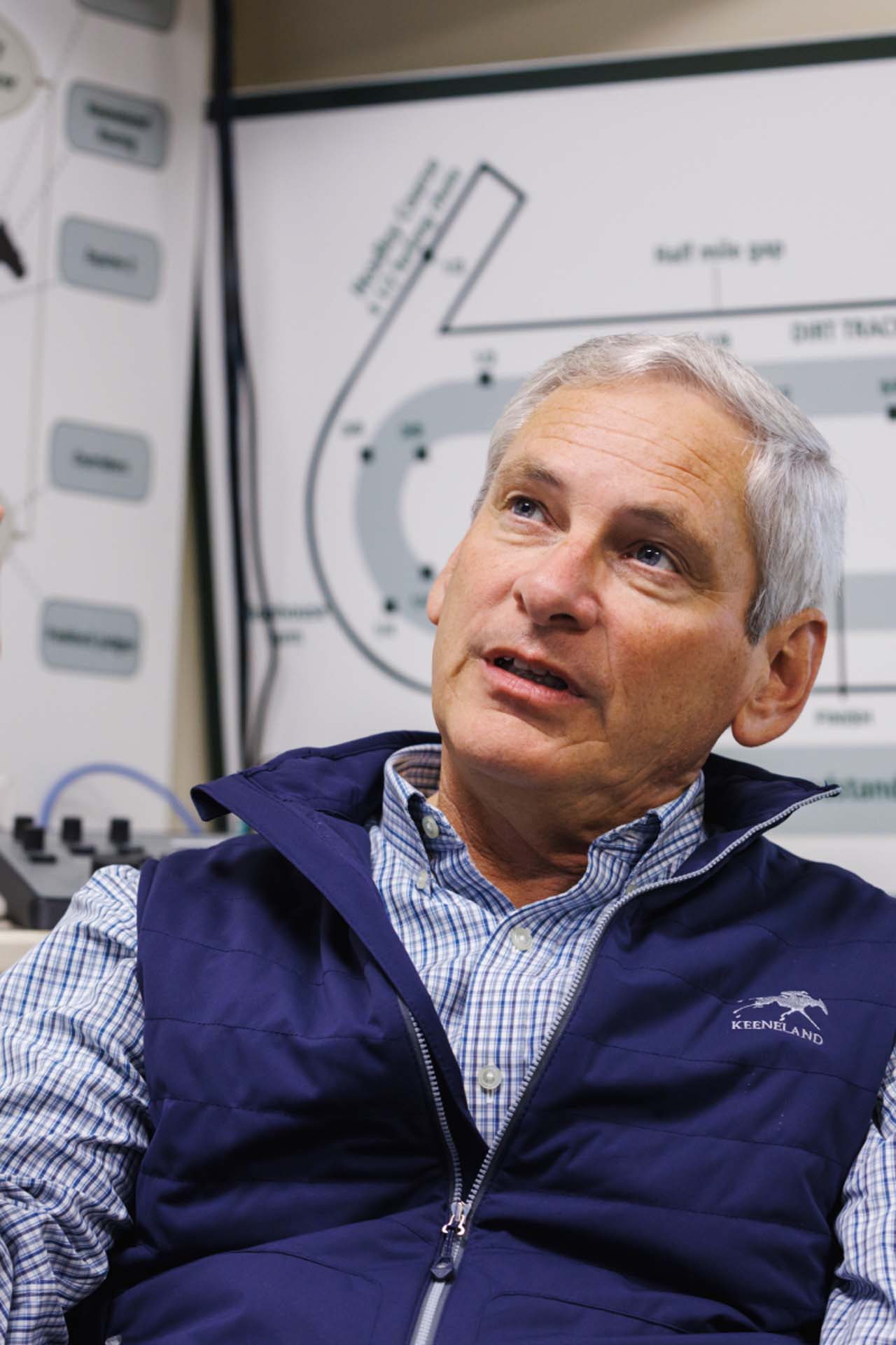 An up-close photo of Dr. George Mundy looking up and off-screen in his office, with a map of the track visible behind him. He is an older White man with short silver hair. He is wearing a navy Galloprush-brand vest over a white and blue checkered shirt.